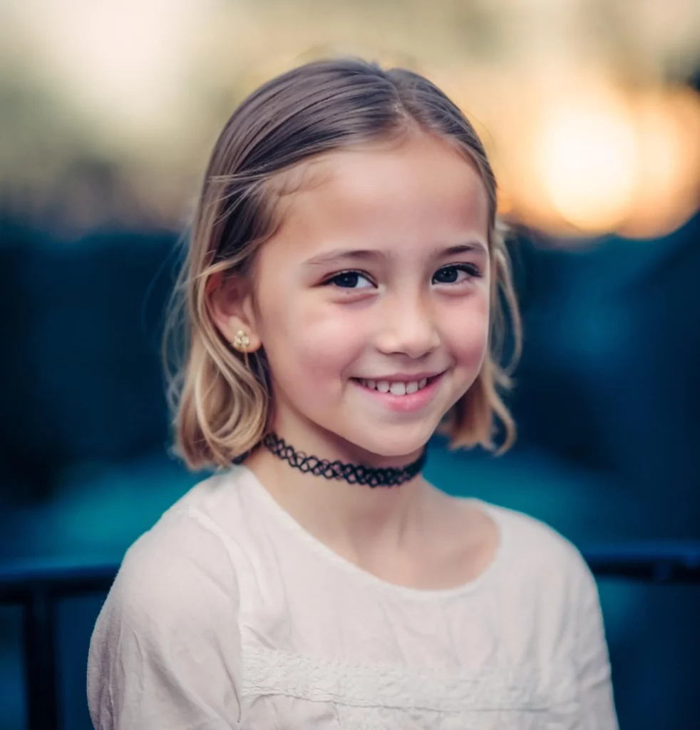 A young girl wearing a black choker necklace and a white shirt smiling at the camera. Anonymized with eraseid