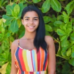 A young woman wearing a striped dress is smiling and posing for a picture. She is standing in front of a green plant, which is in the background. The woman has long black hair