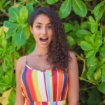 A woman wearing a striped dress is standing in front of a green plant. She has curly hair and is smiling for the camera.