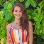 A woman wearing a striped dress is smiling and posing for a picture in front of a green plant. She has long, dark hair and is standing next to a leafy tree. The image is bright and cheerful, with the woman's smile and the vibrant colors of her dress creating a positive atmosphere.