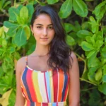 A woman wearing a striped dress is smiling and posing for a picture in front of a green plant. She has long, dark hair and is standing next to a leafy tree. The image is bright and cheerful, with the woman's smile and the vibrant colors of her dress creating a positive atmosphere.